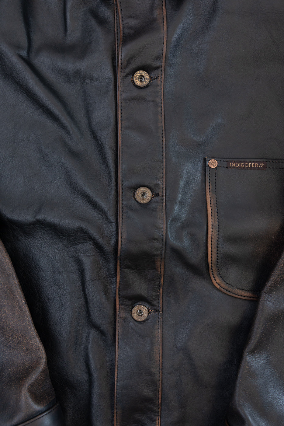 Close-up of the front of a dark brown leather shirt showing buttons, texture, and pocket tag.