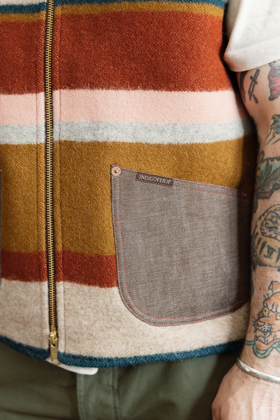 Close-up front view of striped wool vest with INDIGOFERA pocket, gold zipper, and red stitching, worn by male model.
