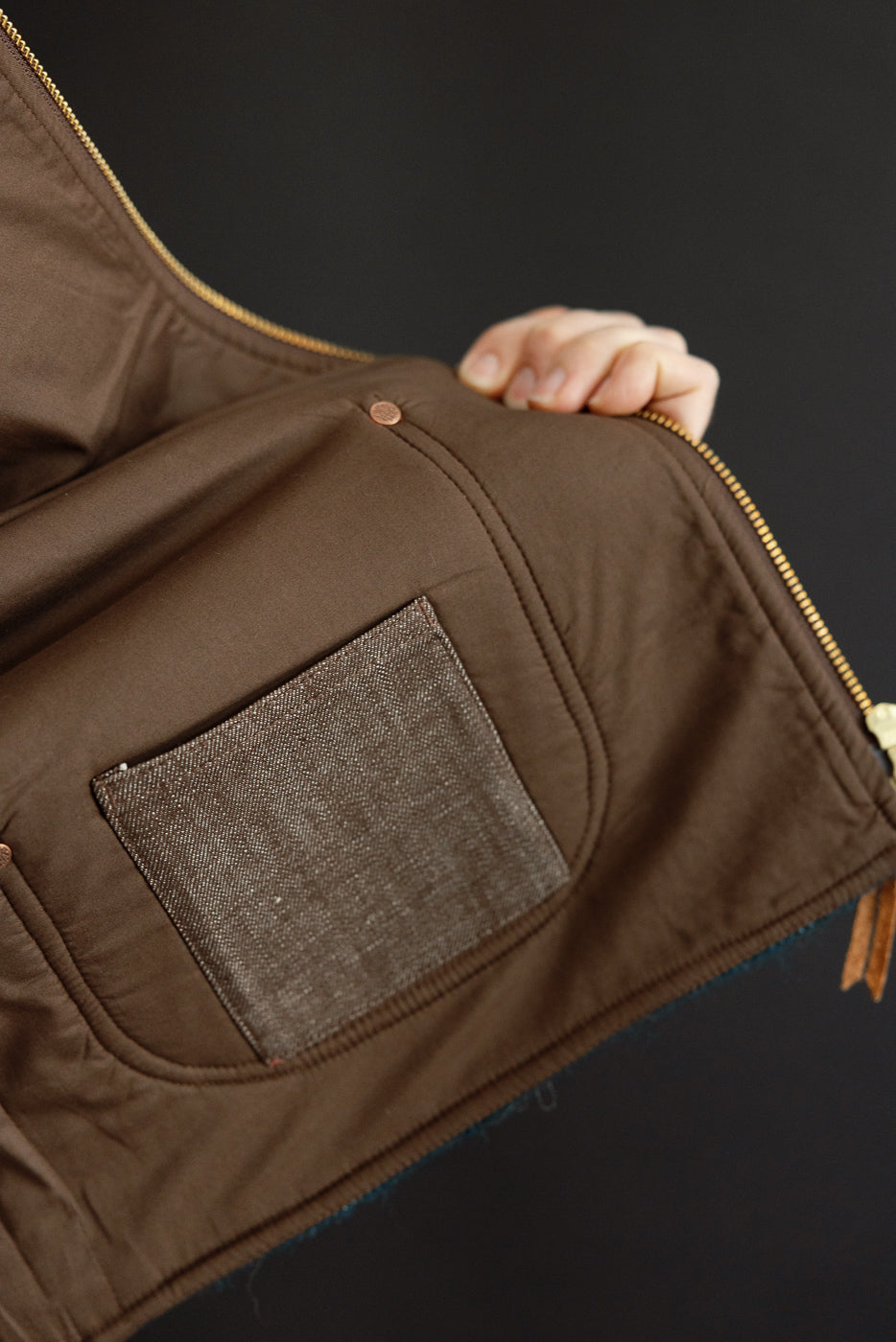 Close-up of brown jacket's front left pocket with textured patch, copper snap, and gold zipper detail