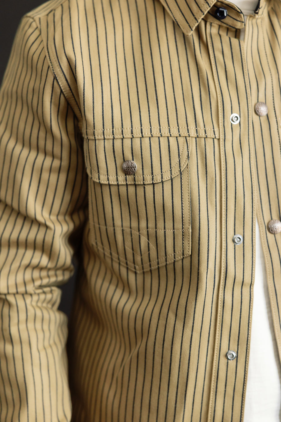 Close-up of male model's front left chest in beige and indigo striped shirt-jack, showing snap button pockets, pointed collar, and shirt texture. Male model is 5'9