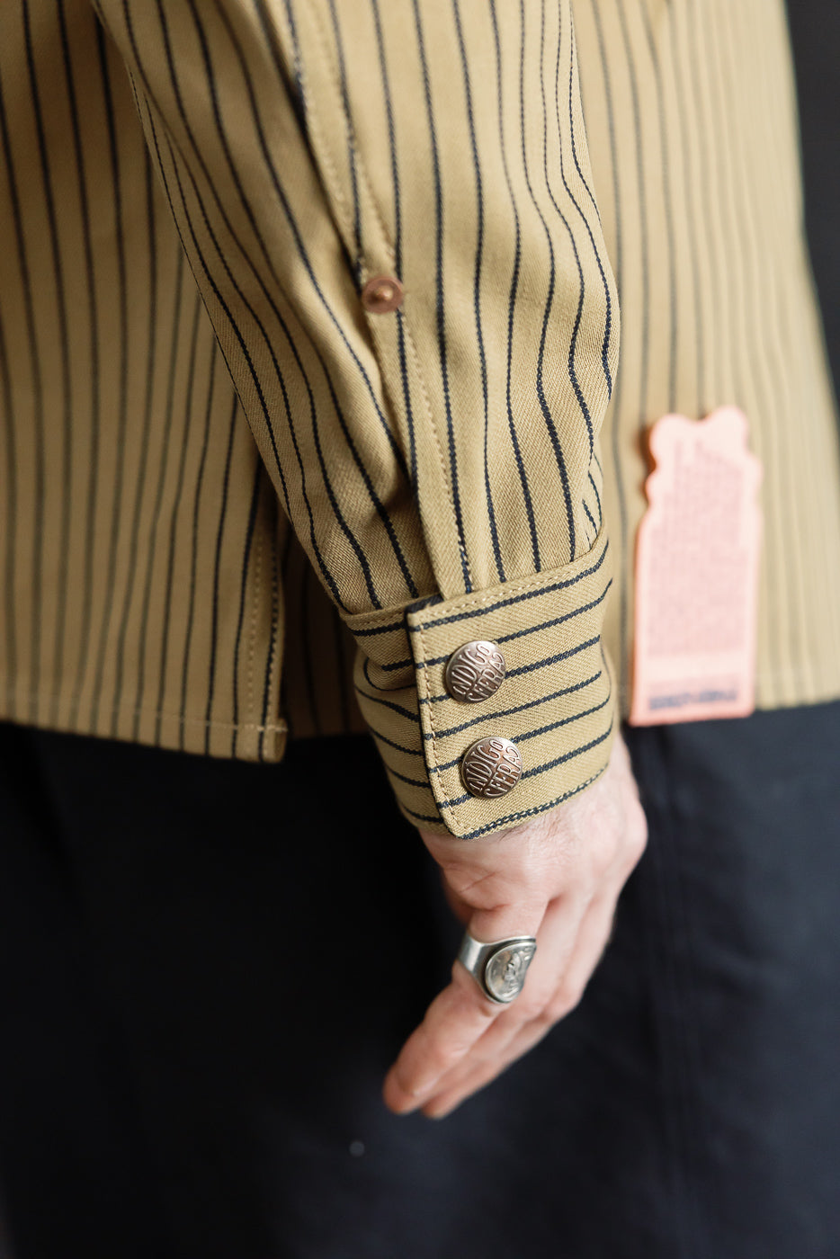 Close-up of male model's right shirt cuff, beige indigo striped fabric, metal buttons, side view.