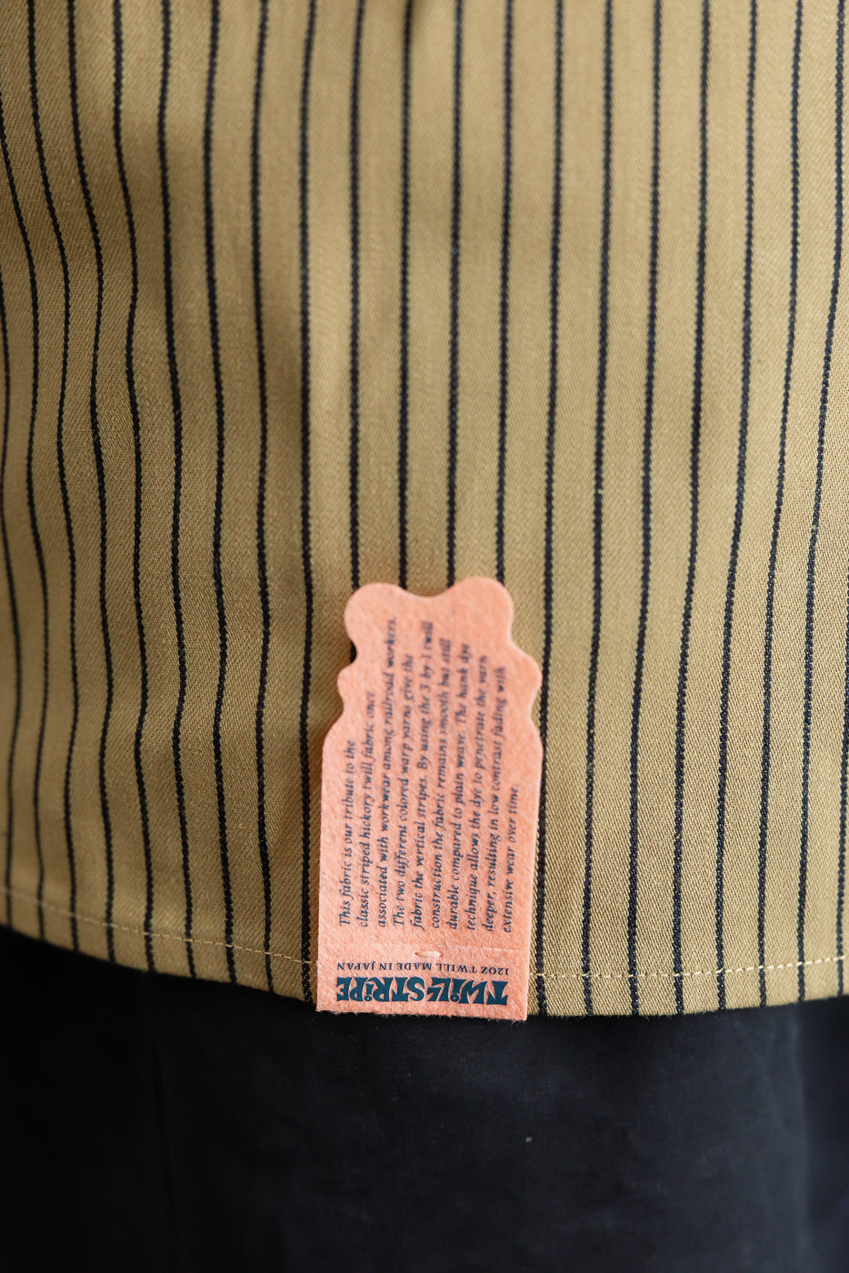 Close-up of lower front hem on beige indigo striped shirt showing Twill Stripe fabric tag detail.