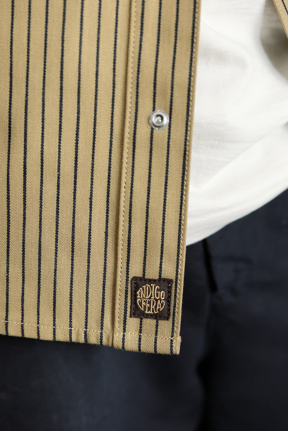 Close-up of male model's left front hem, showing beige indigo striped shirt fabric, snap button, and woven tag detail.