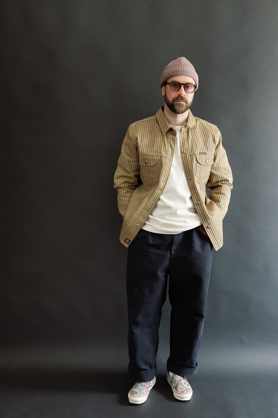 Full body front view of male model, 5'9'', 165lbs, athletic build, wearing beige indigo striped shirt jacket, white t-shirt, dark pants, and patterned slip-on shoes.