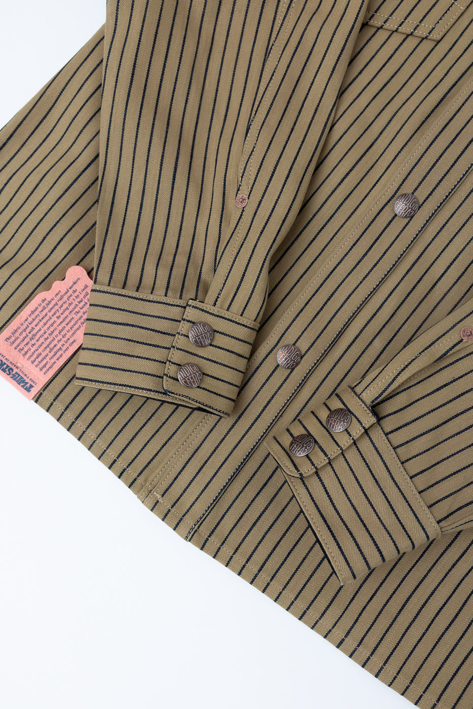 Close-up of shirt jacket front, beige with indigo stripes, showing cuff, buttons, fabric texture, and tag.