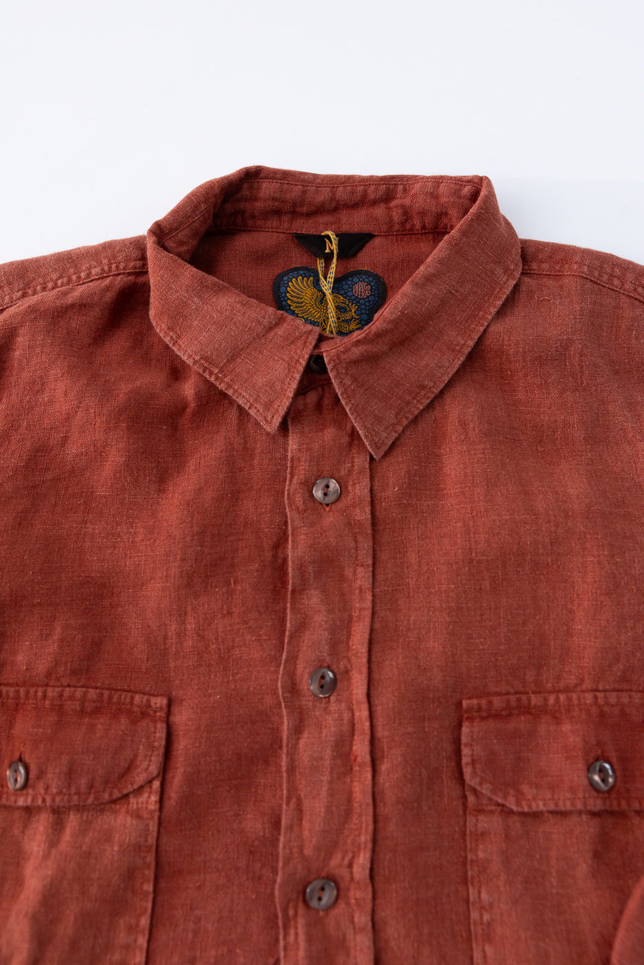 Close-up front view of rust linen shirt with button details, collar, and tag, showcasing texture and design.