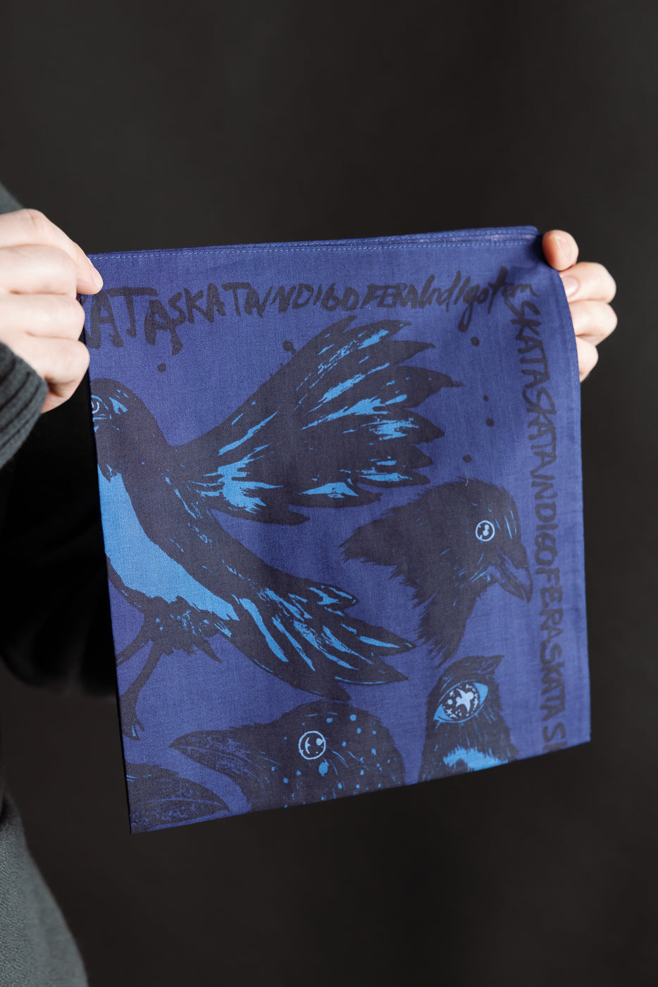 Close-up front view of Indigofera Devils Bird bandana in blue and black with bird graphic and edge logo, held by hands. Contemporary casual accessory at Dant.