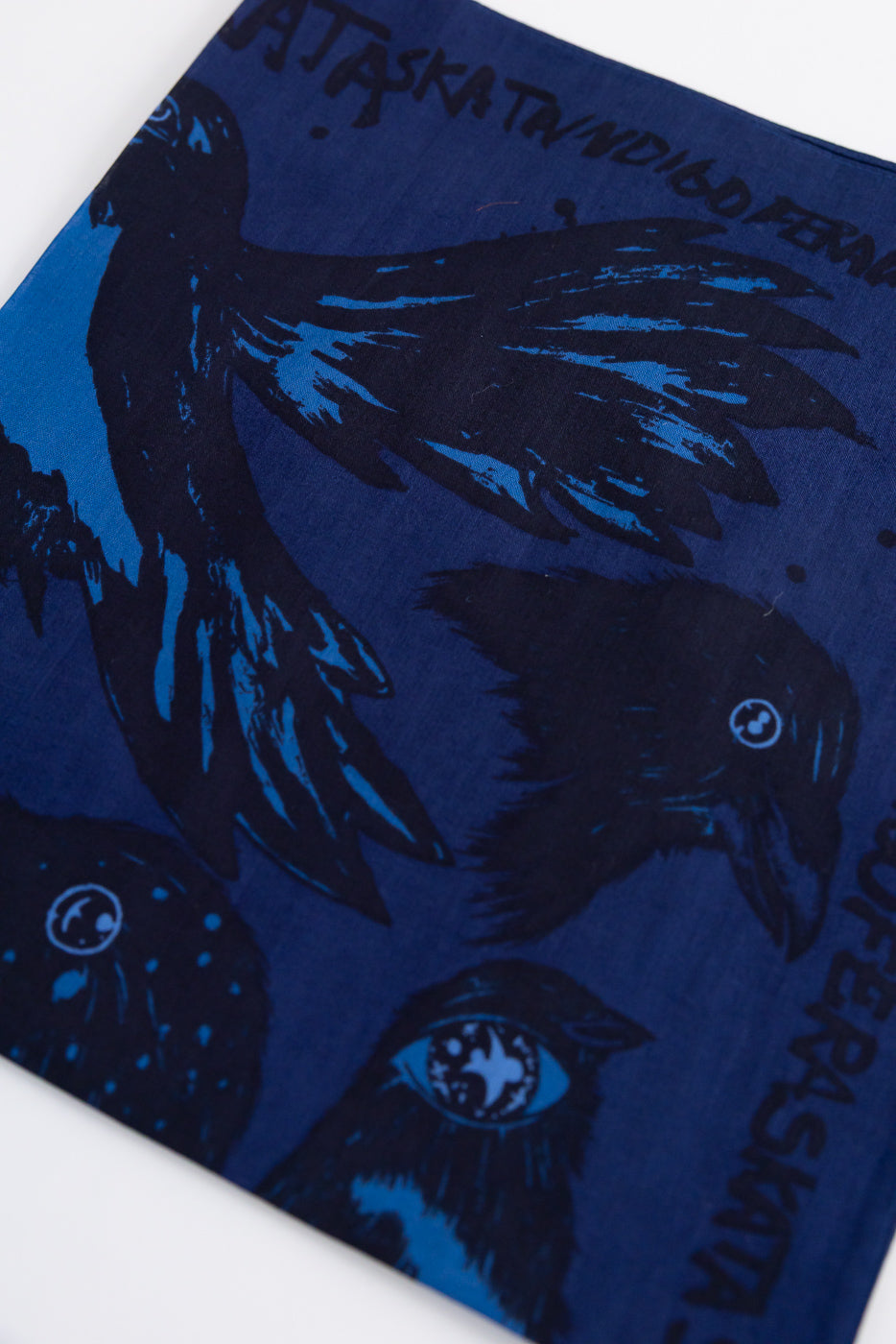 Close-up of Indigofera x Ragnar Persson blue-black bandana, front side with crow artwork, contemporary casual accessory at Dant.