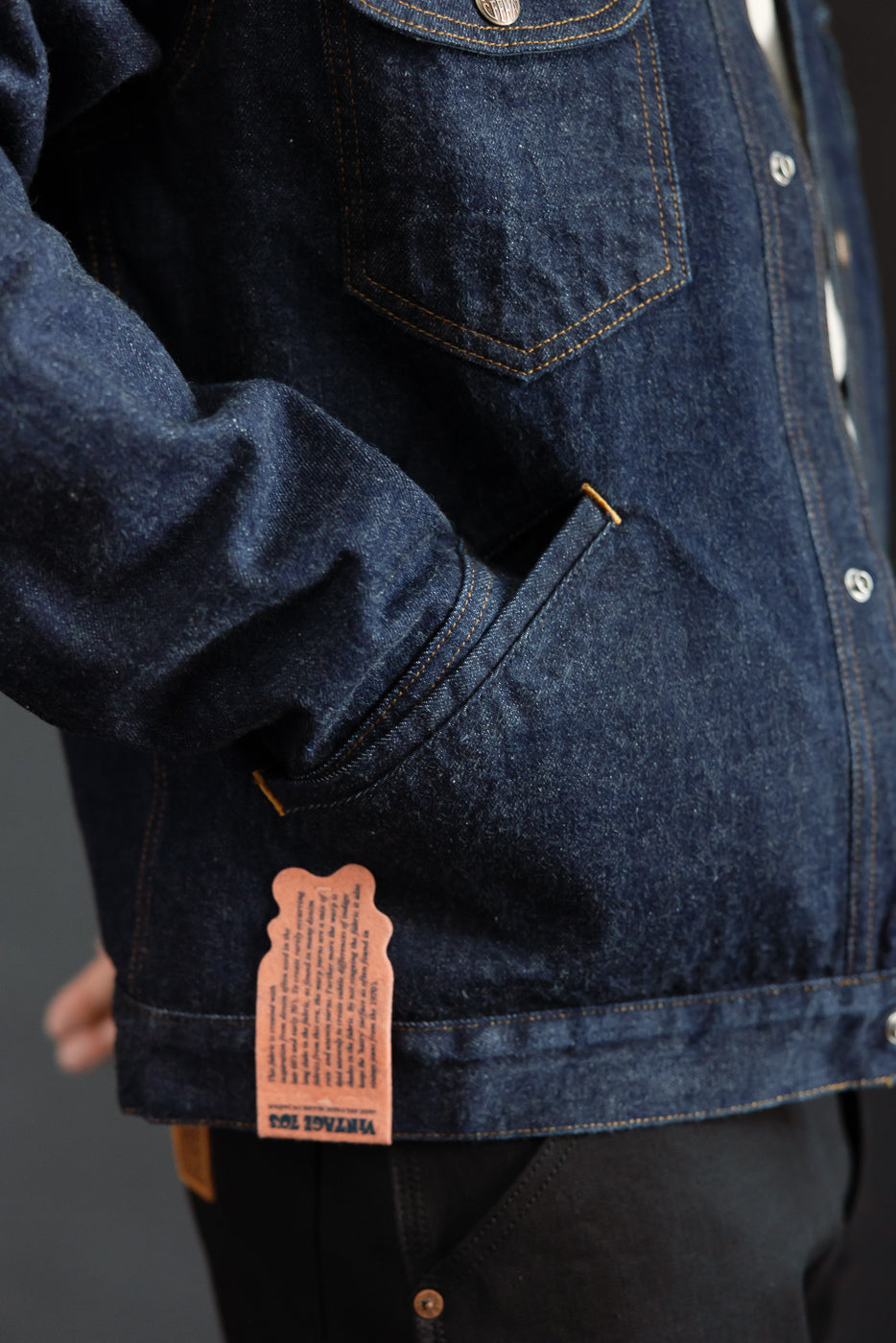 Close-up of male model's side view showing dark denim jacket with orange stitching, front pocket, and leather tag.