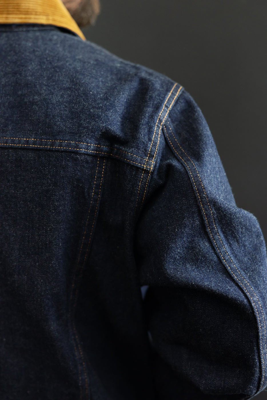 Close-up of male model's back shoulder, showcasing denim jacket with contrast stitching and corduroy collar.