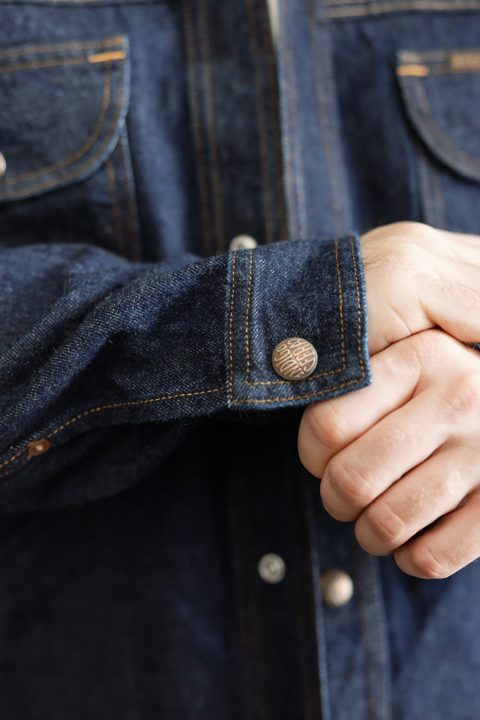 Close-up of male model's denim shirt sleeve cuff, showcasing button detail and fabric texture.