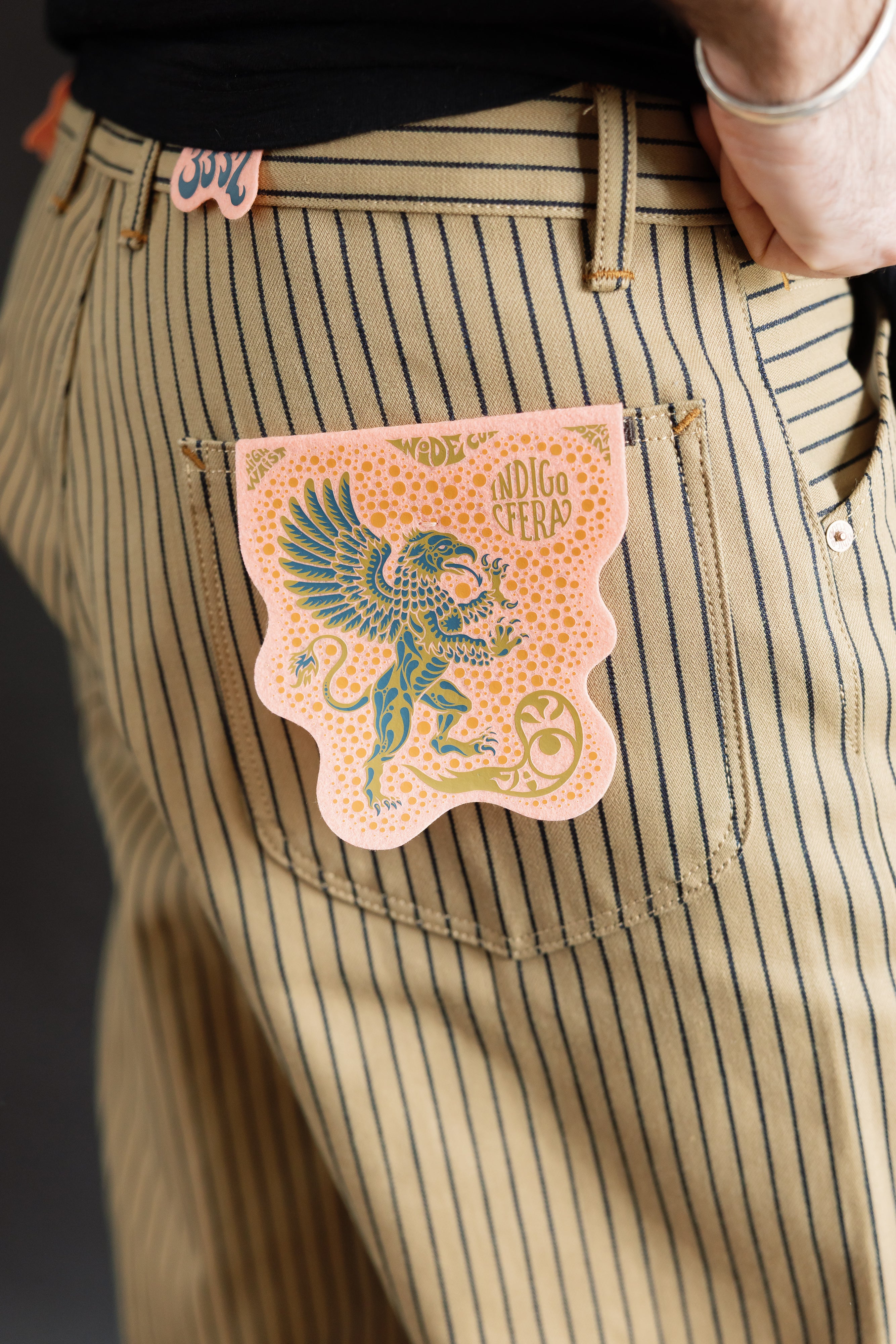 Close-up of male model's right hip wearing beige indigo striped pants, showing back pocket and paper patch detail. Model is 5'9