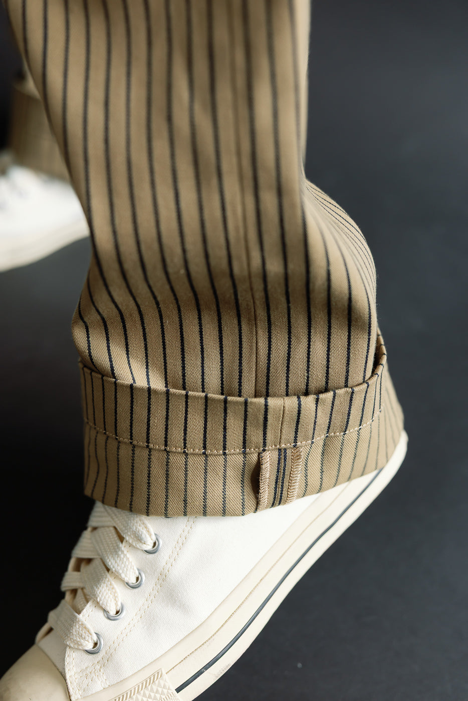 Close-up of male model's lower leg, showcasing beige indigo striped pants cuff and sneaker, side view.