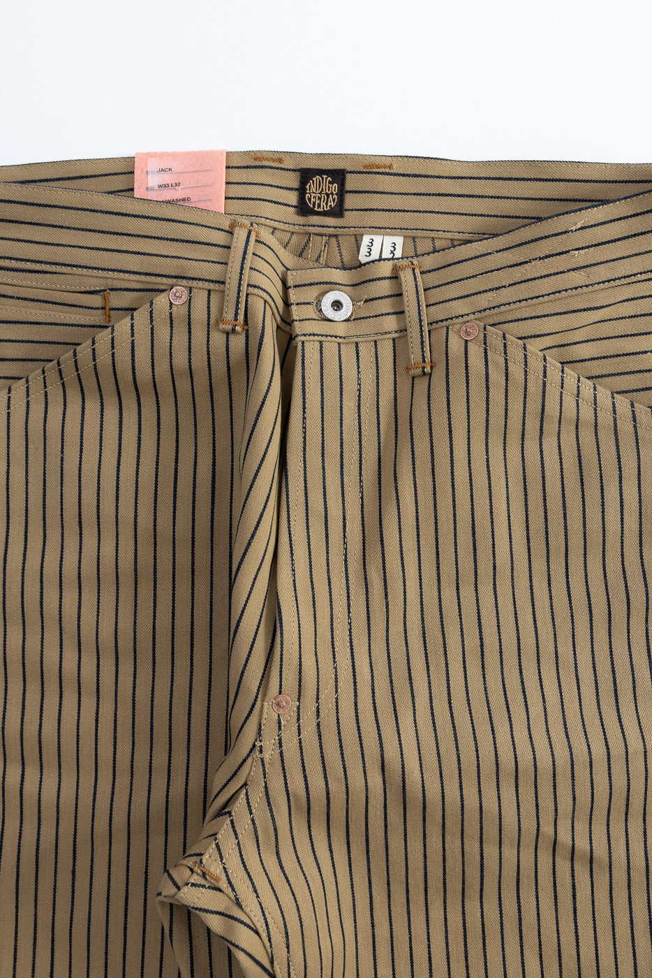 Close-up of front waist and fly area on beige and indigo striped pants, showing button, belt loops, and tags.