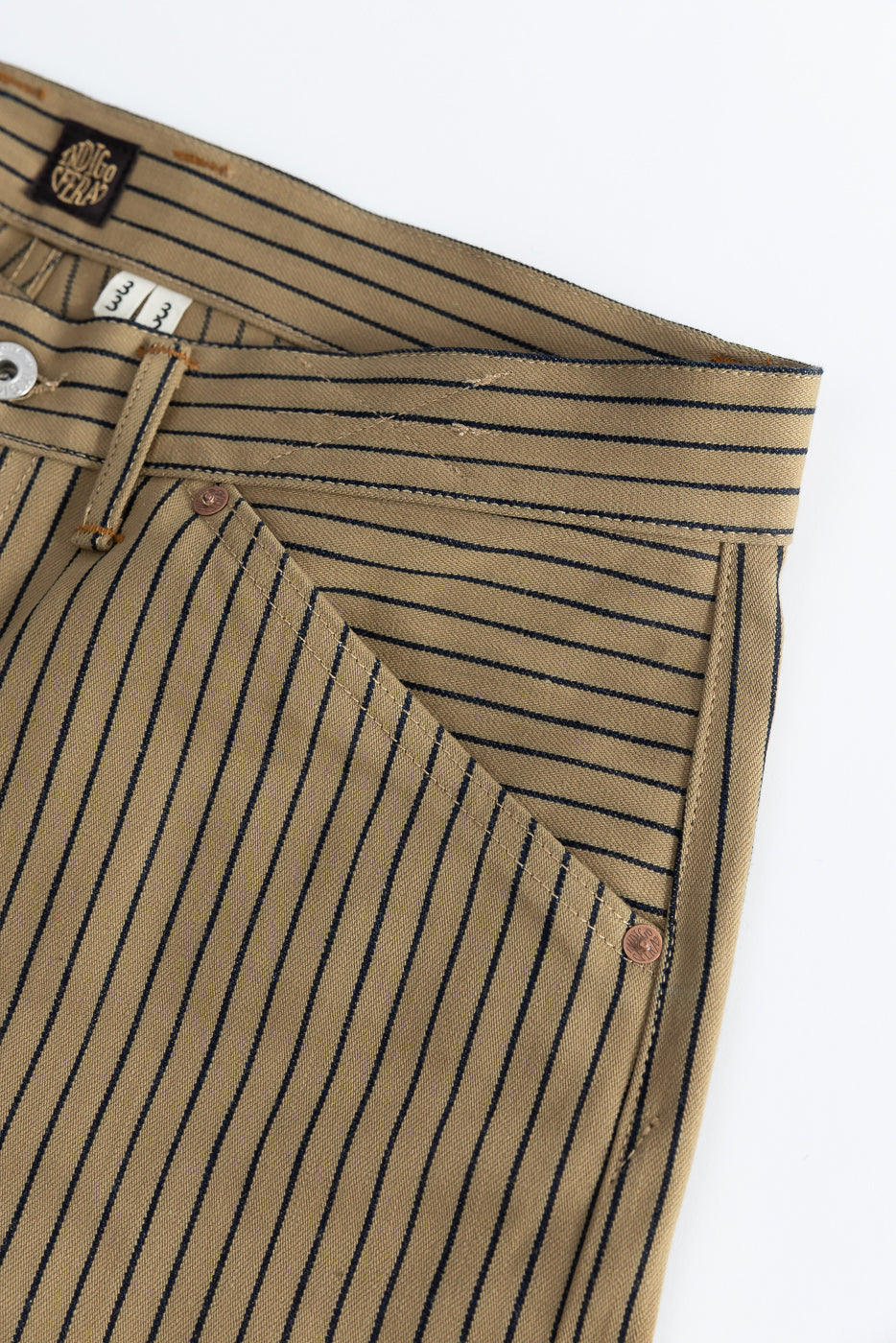 Close-up of front left hip and pocket on beige indigo striped pants, showing fabric texture, rivet, belt loop, and branded tag.