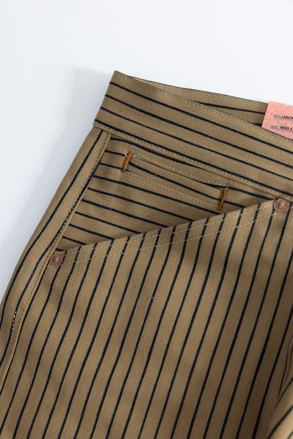 Close-up of front waist and pocket on beige and indigo striped men's pants, showing fabric texture, stitching details, and rivets.