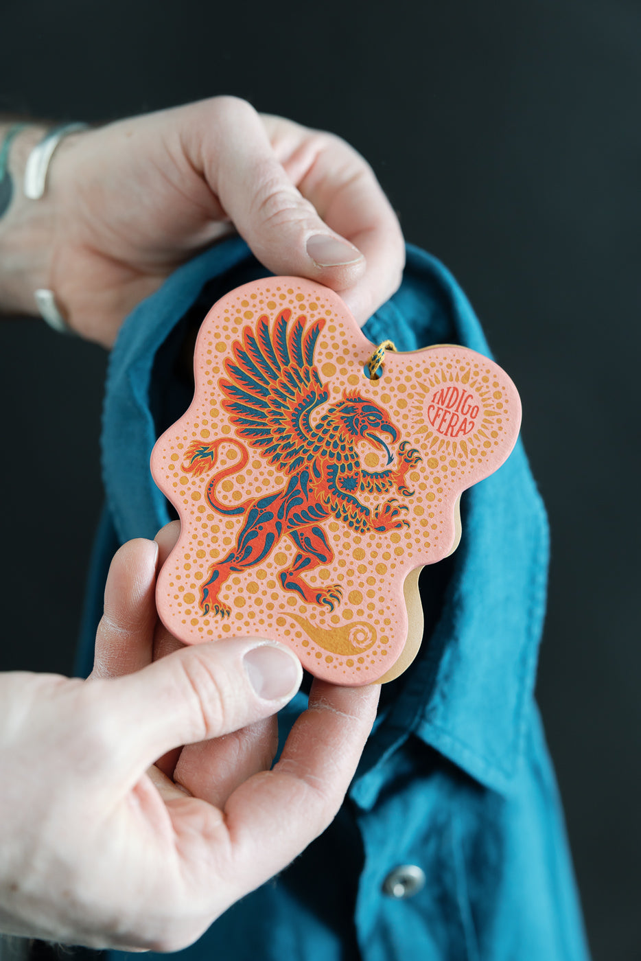 Close-up of a male hand holding an Indigofera brand tag with griffin design, over the front chest of an ocean blue canvas shirt, highlighting fabric texture and snap button detail.