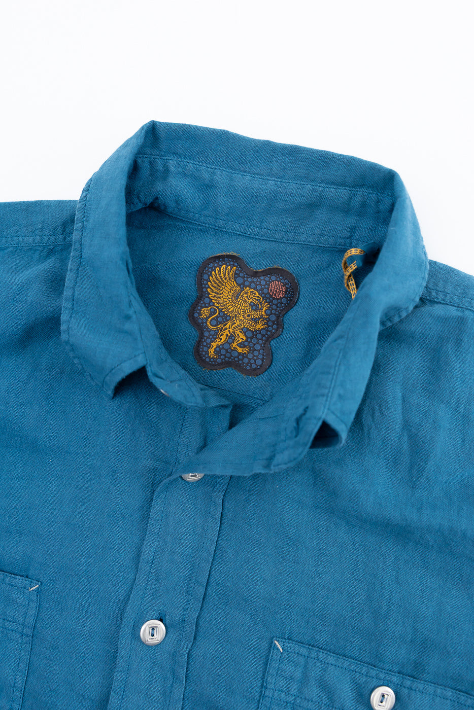 Close-up of Indigofera McAllen shirt front, ocean blue canvas, showing collar, fabric texture, buttons, and embroidered inner neck tag.