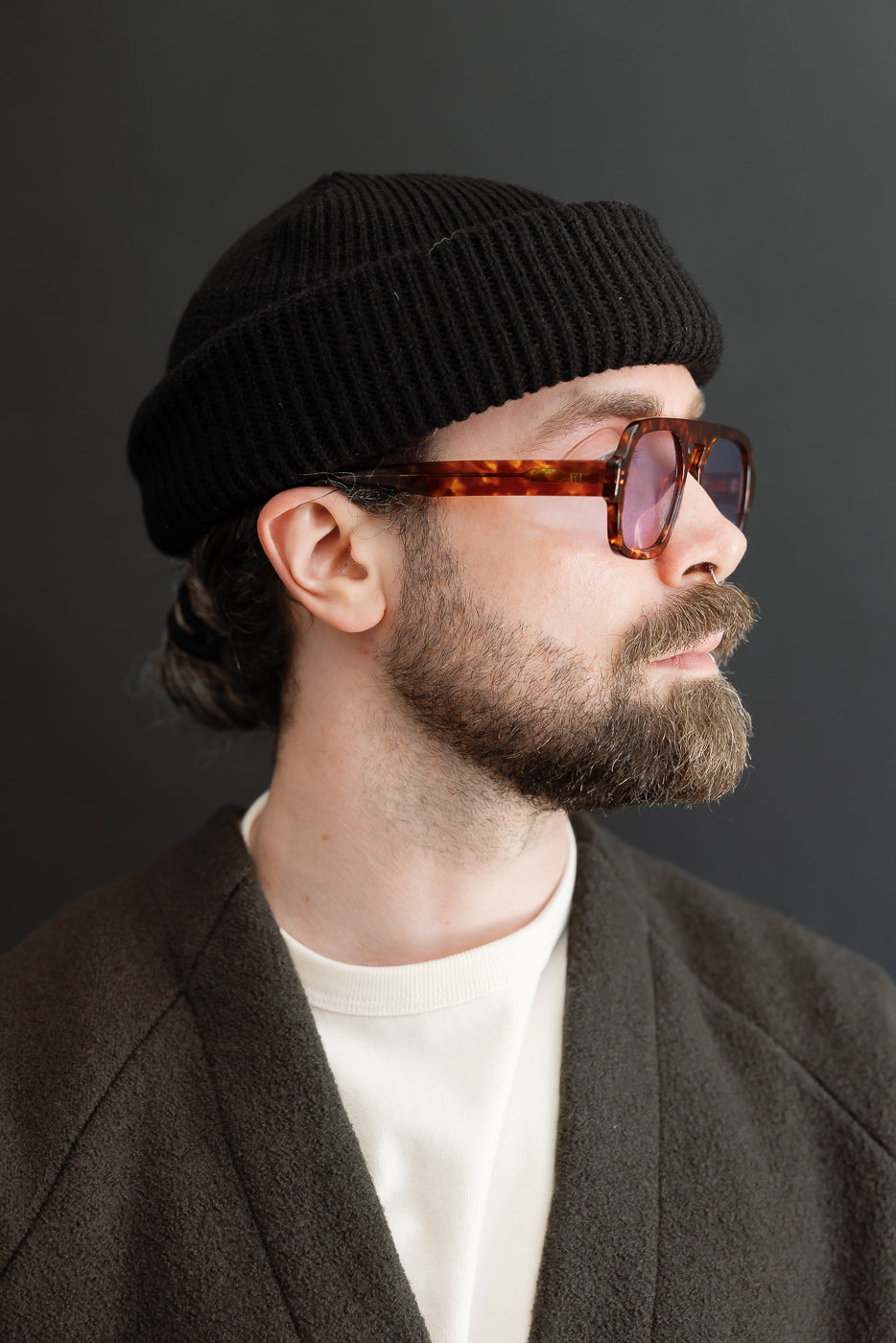 Close-up side profile of a male model wearing tortoiseshell sunglasses, a black beanie, and a brown jacket.