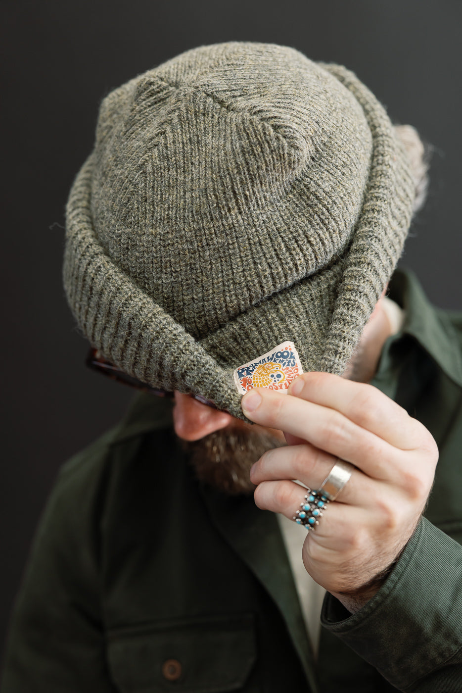 Close-up front view of a male model wearing a textured olive green knit beanie with a small tag.