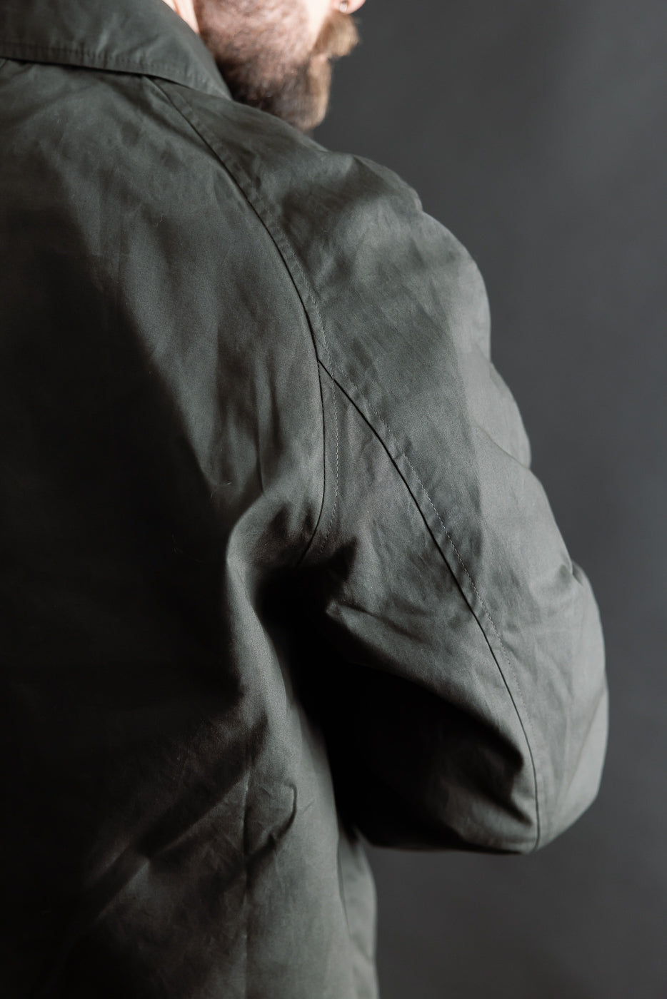 Close-up of male model shoulder and sleeve, Indigofera Raven Coat dark grey coated cotton twill, left rear side, showing fabric texture and seam detail. Model is 5'9