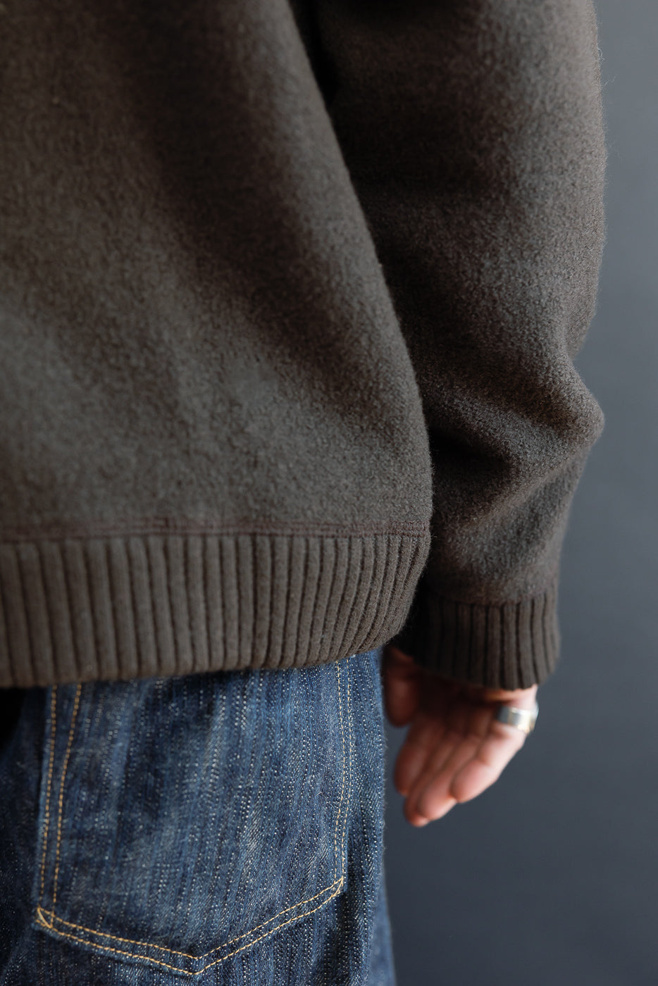 Close-up of the back hem and side view of a male model wearing a brown textured sweater and denim jeans.