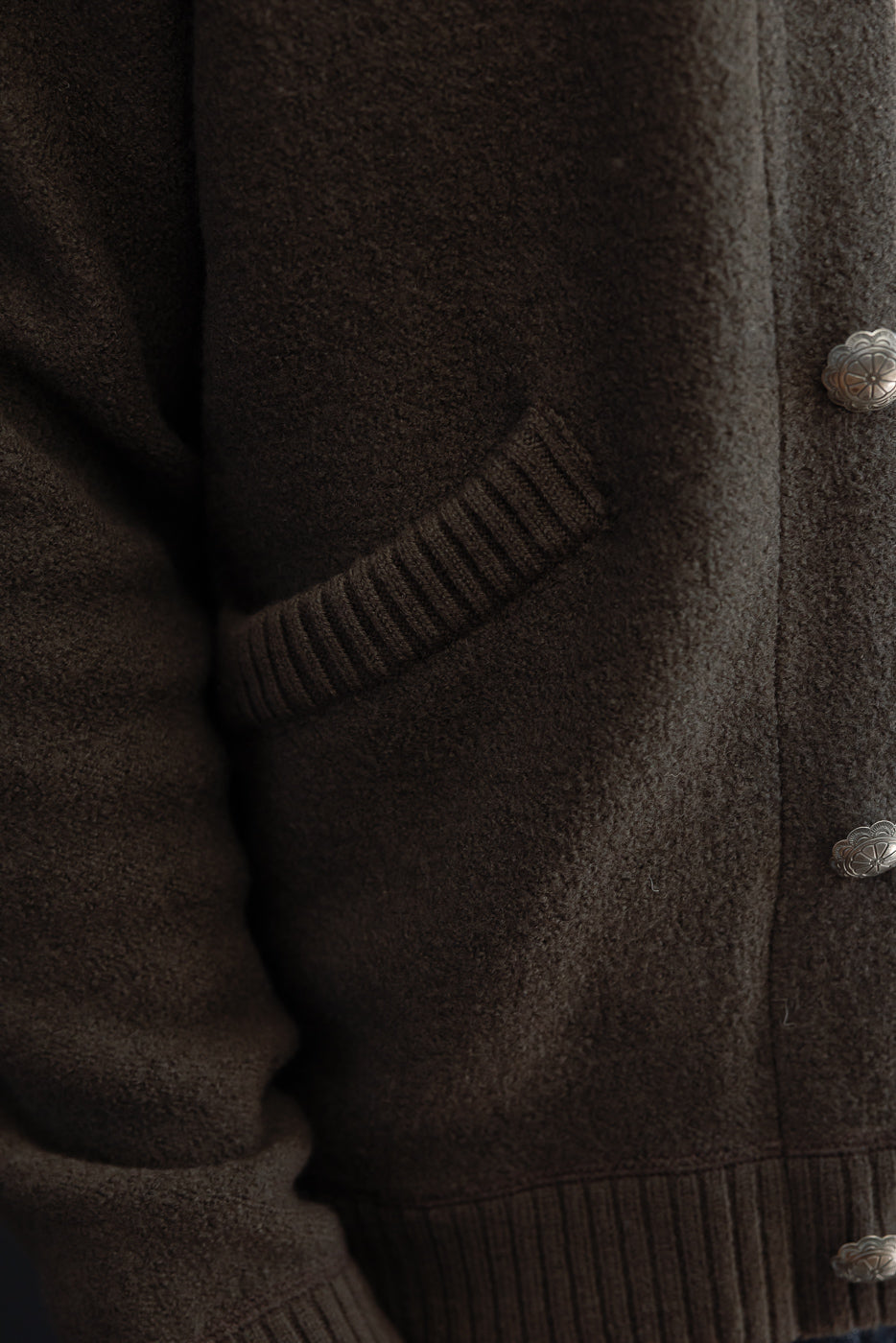 Close-up of the front right side of a brown textured wool cardigan showing a ribbed pocket and metal buttons.