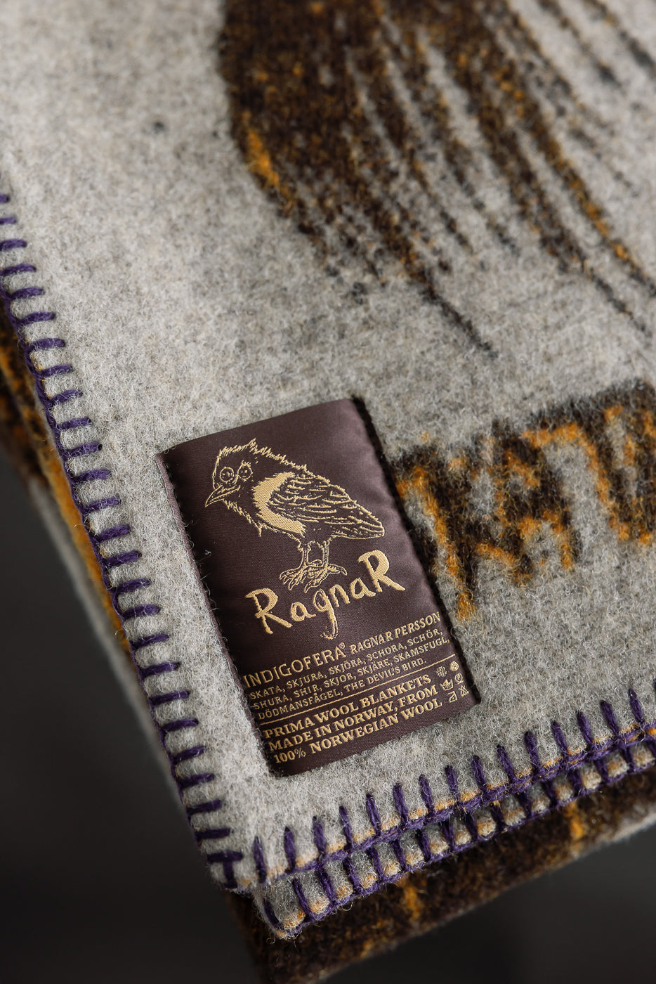 Close-up of Indigofera Skata Blanket grey brown yellow edge with Ragnar Persson collab tag, whipstitch hem, 100% Norwegian wool