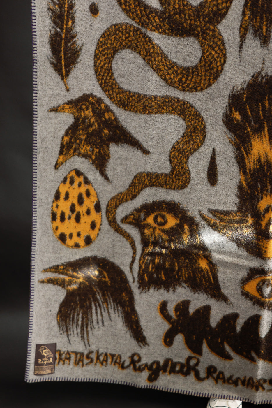 Close-up of Indigofera Skata blanket front corner, grey with brown and yellow bird, egg, and snake graphic details, visible tag and stitched edge, Dant contemporary casual accessory.