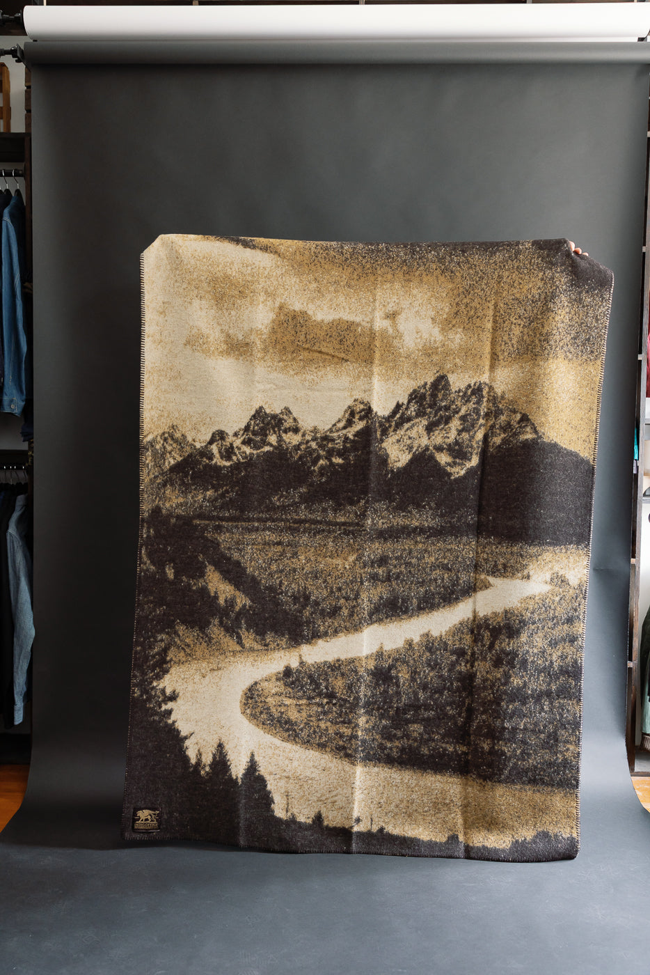 full image of snake river blanket being held up. landscape painting with beige and brown skies, mountain range, and a prairie featuring the snake river. all the details are in black and beige. front view
