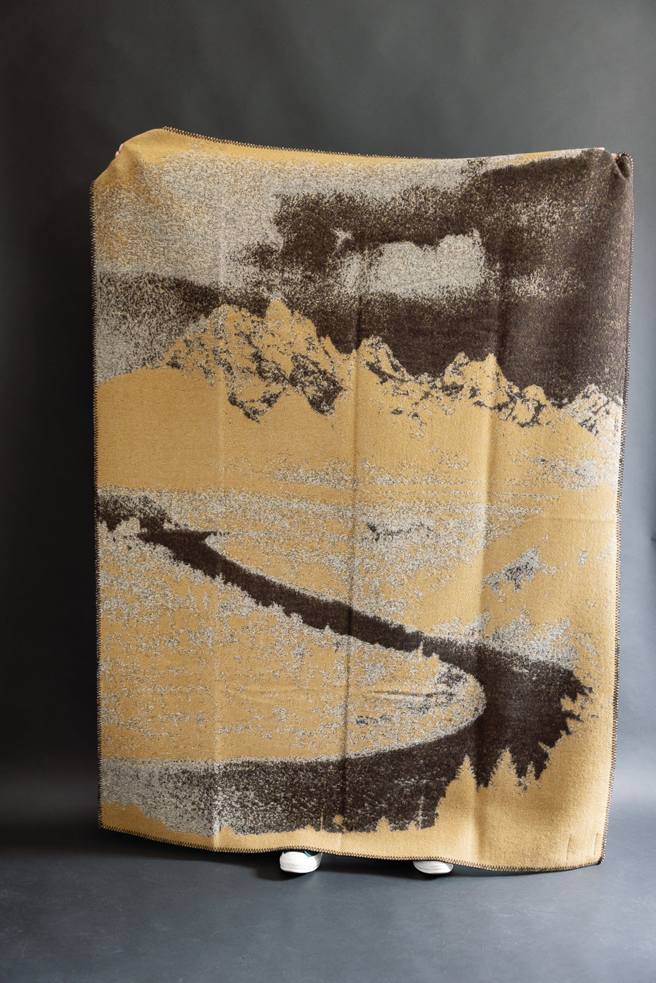 full image of snake river blanket being held up. landscape painting with beige and brown skies, mountain range, and a prairie featuring the snake river. all the details are in black and beige. reverse image, the negative of the front