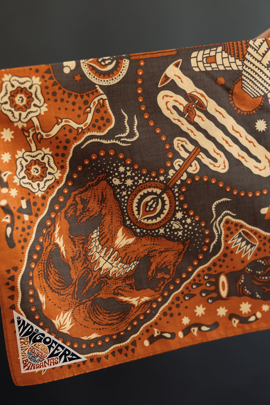 Close-up of front of orange and brown bandana with skull, floral, and geometric patterns, featuring Indigofera tag.