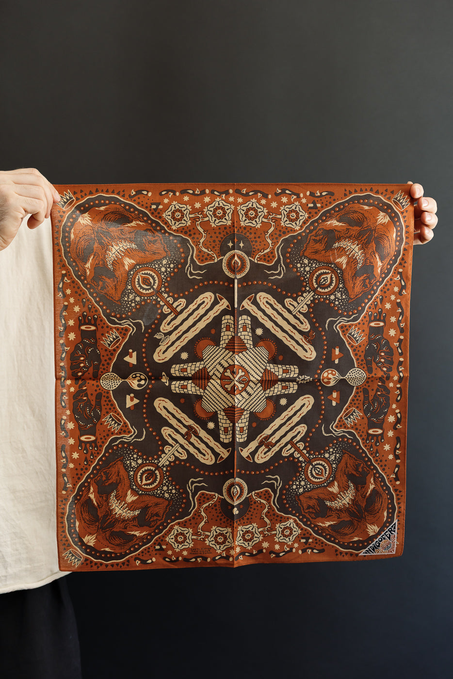 Male model holding a detailed brown bandana with skull and geometric patterns, front view, full design visible.