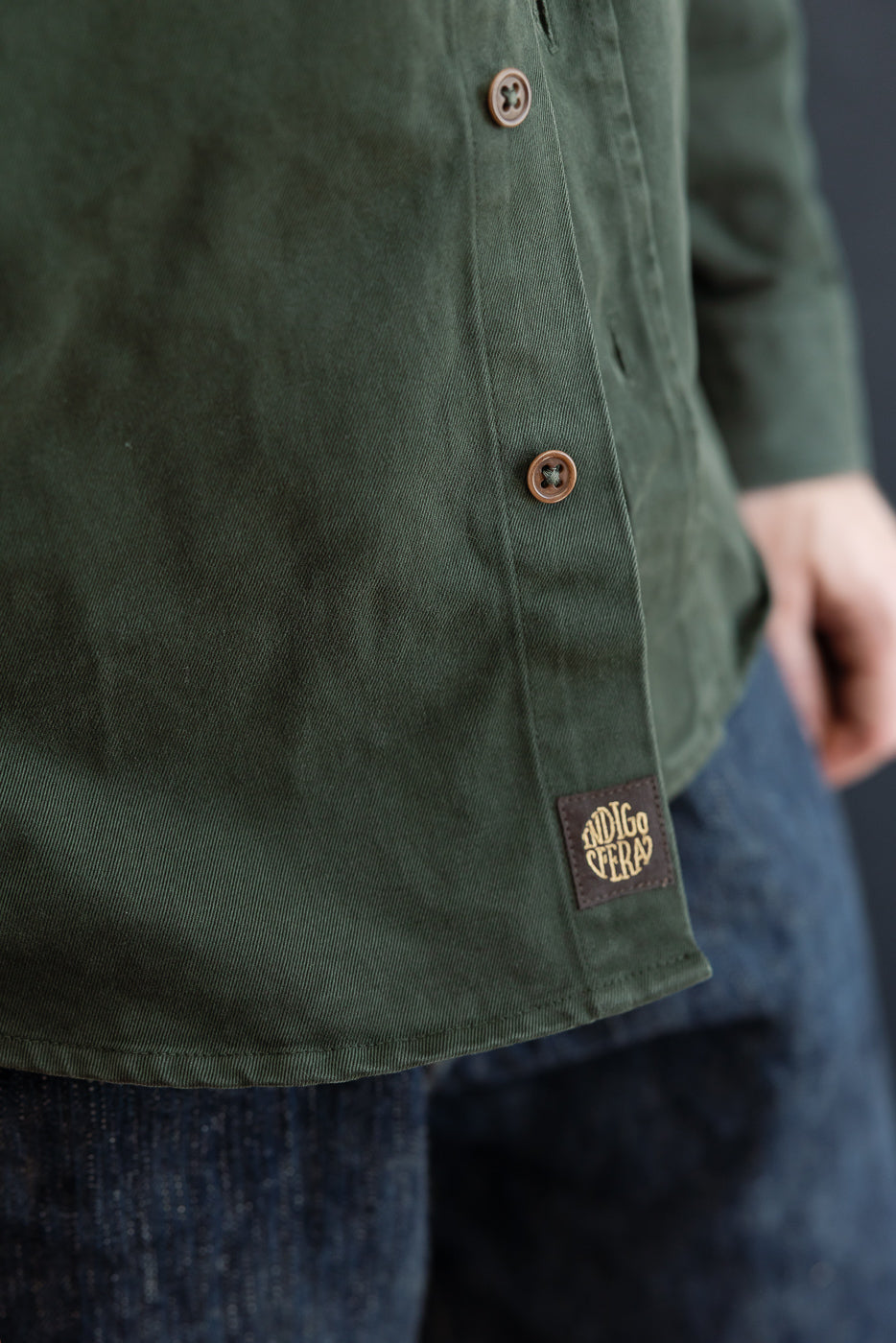 Close-up of the front bottom hem of a male model's green cotton shirt, showing a brown Indigofera tag.