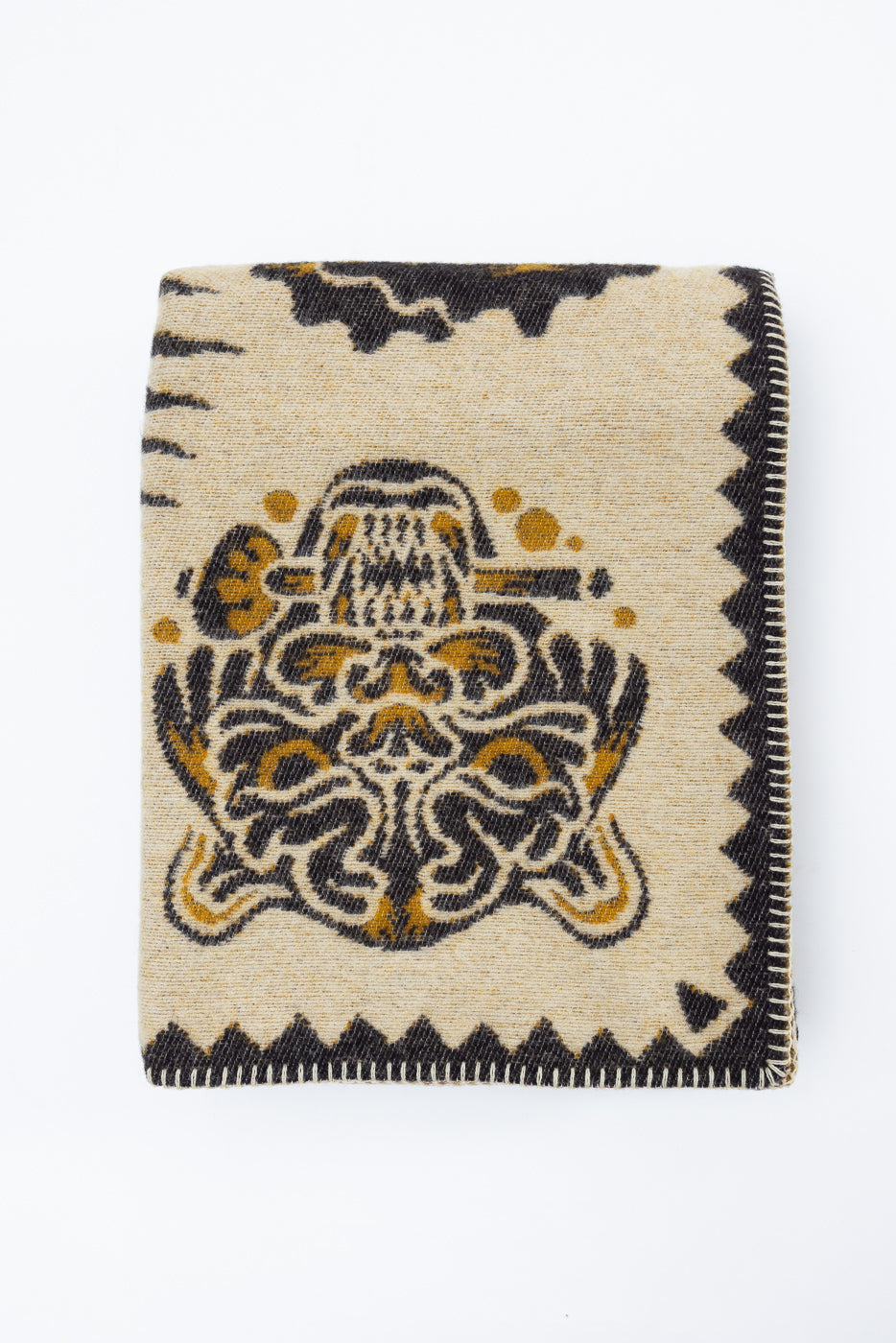 Flat lay, front view of a folded tan knit scarf with an intricate black and gold graphic design.
