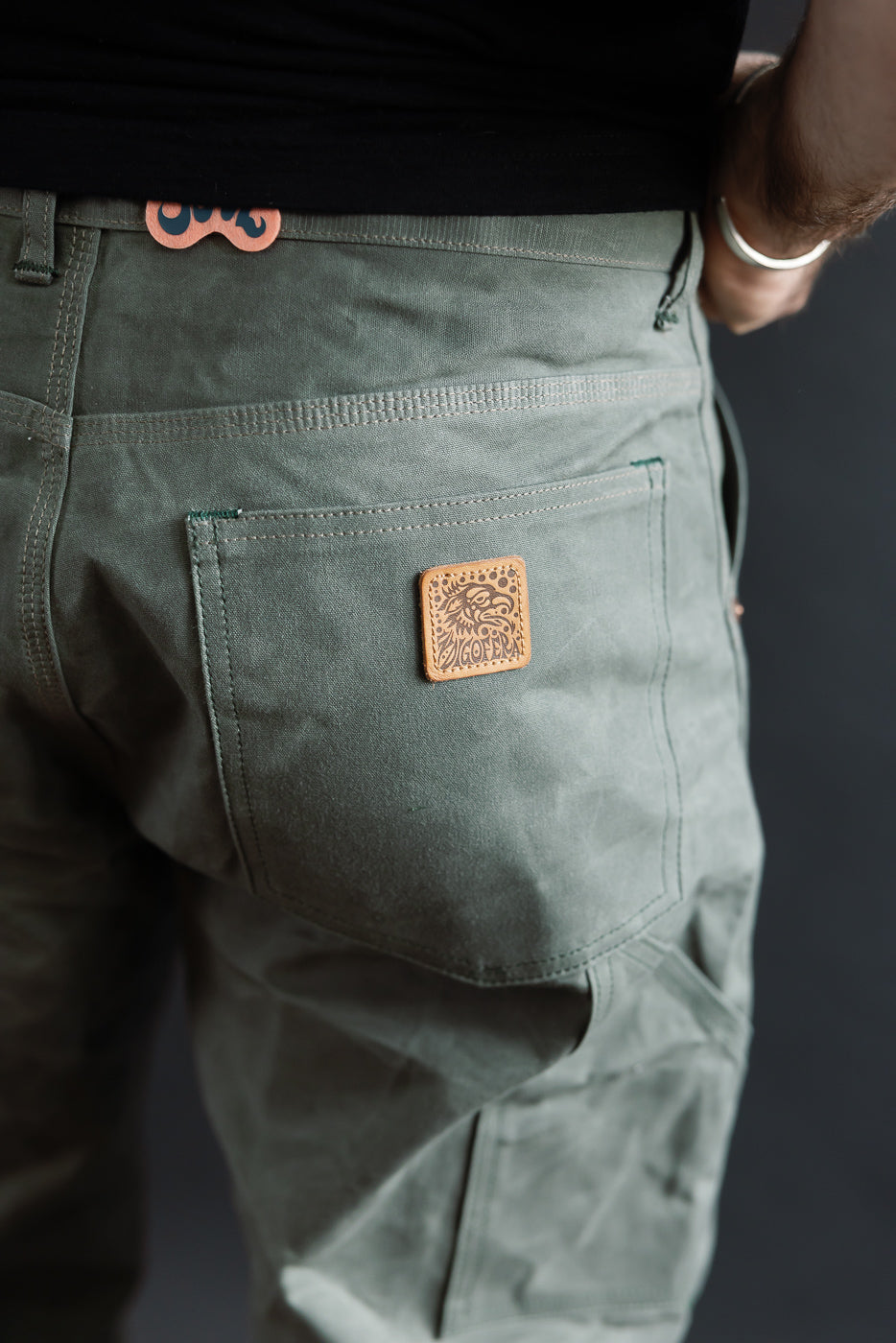 Close-up of rear side of greyish coated canvas work pants on male model, highlighting back pocket and leather logo patch. Model is 5'9