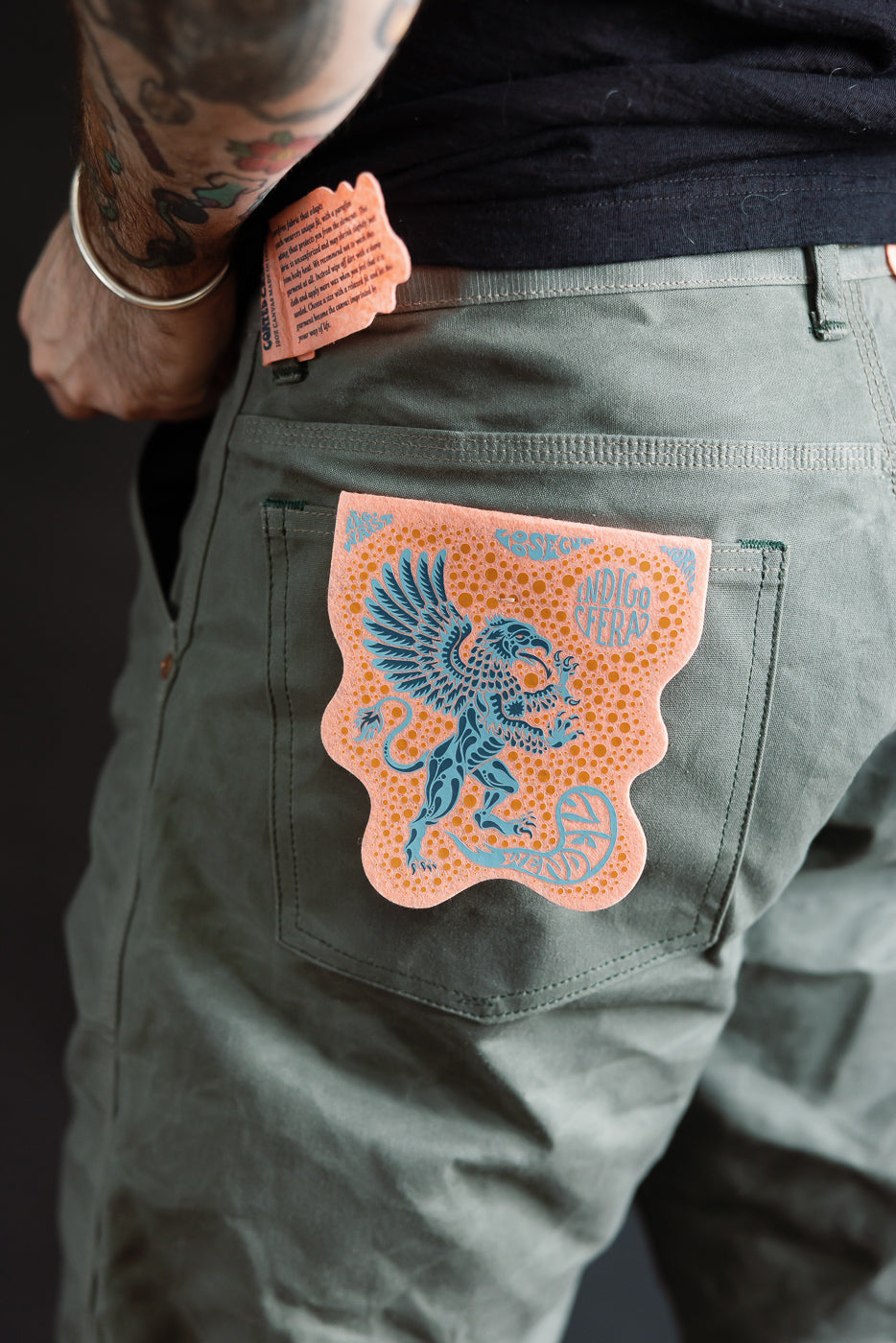 Close-up of back right side on male model, 5'9”, 165lbs, athletic build, showing olive coated canvas pants with orange and blue graphic patch on pocket and visible label detail.