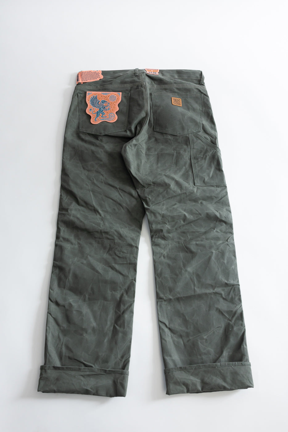 Flat lay of men's coated canvas double knee pants, back view showing two back pockets and detailed leather patch on left pocket.