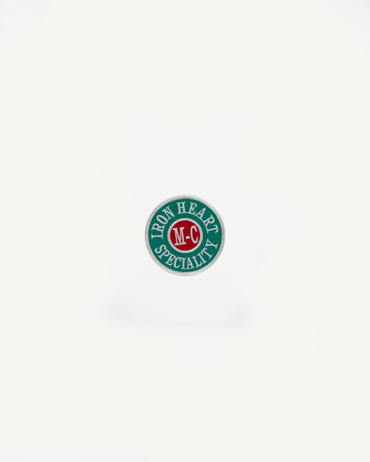 Circular embroidered patch: green background, white 'IRON HEART SPECIALITY' text, red center with 'M-C', white border