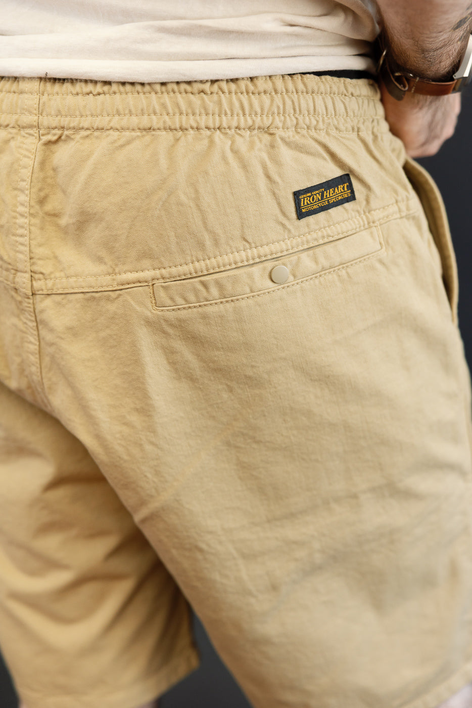 Back close-up of male model wearing beige shorts with Iron Heart tag, buttoned pocket, and elastic waist.