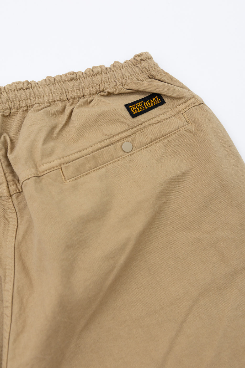Close-up back view of tan chino shorts showing elastic waistband, rear pocket, button, and Iron Heart brand tag