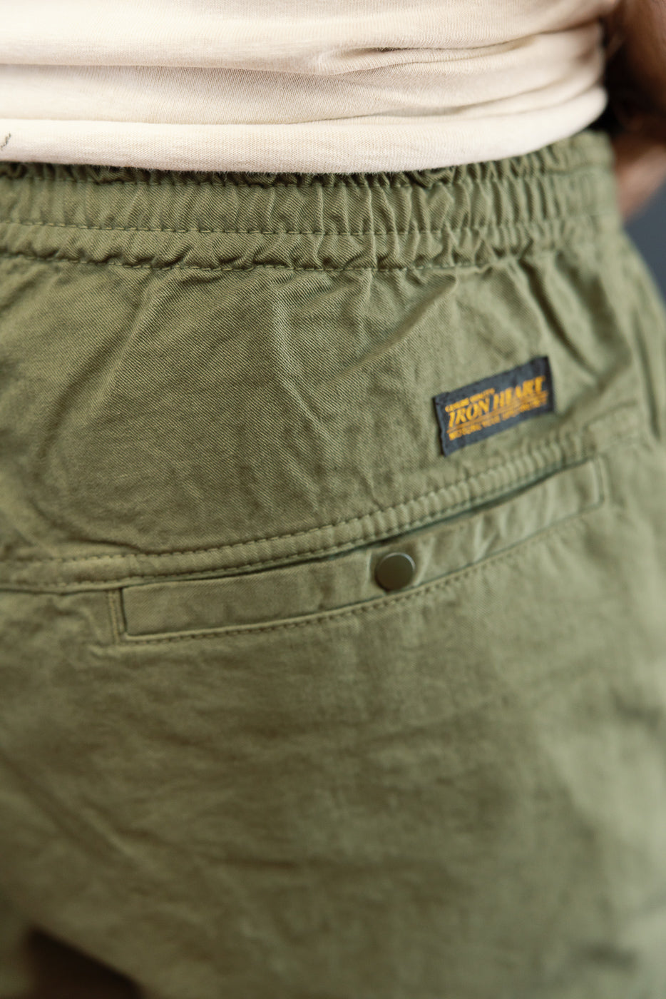Close-up back view of olive green shorts with Iron Heart patch, buttoned pocket, elastic waist, worn by male model
