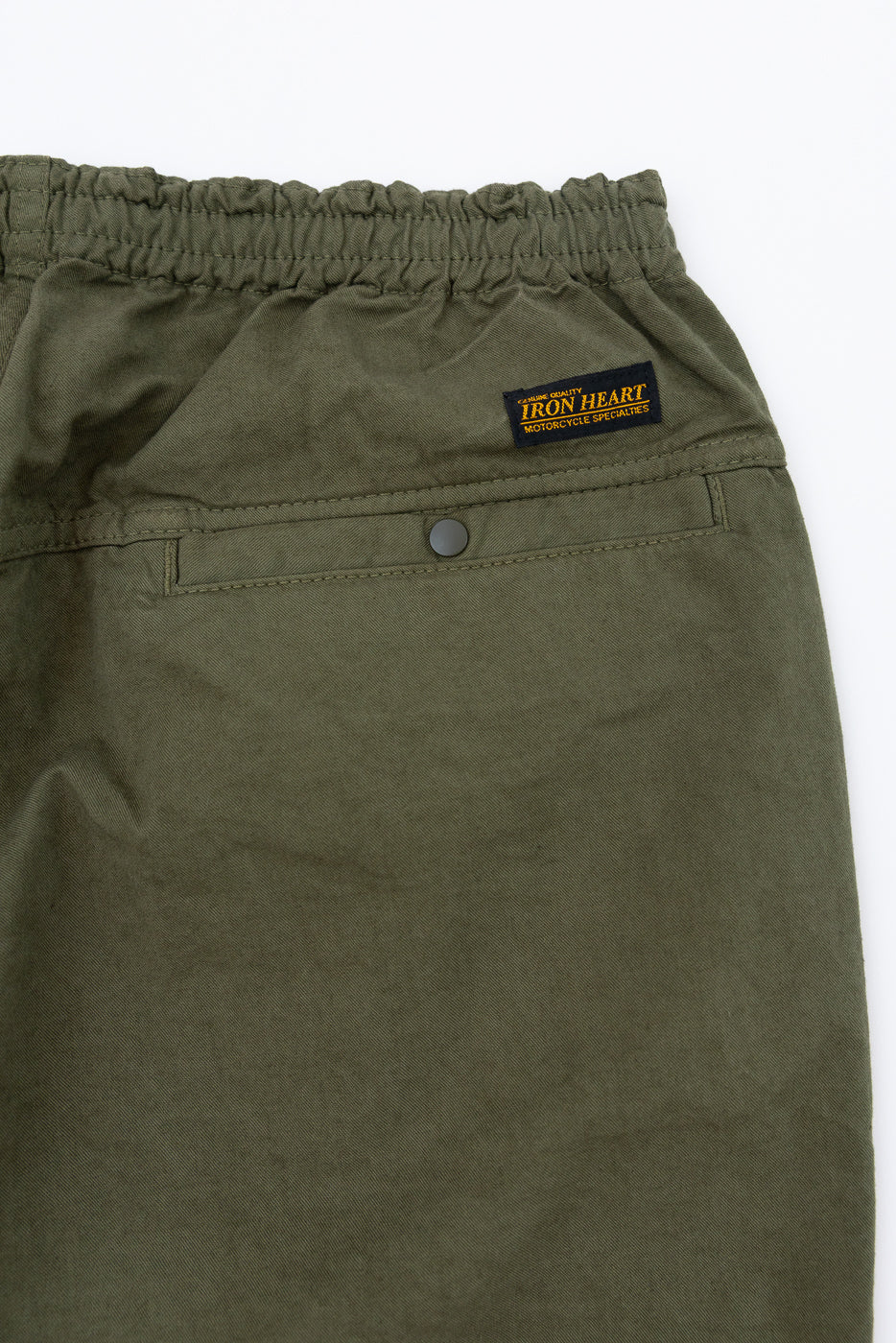 Close-up front view of olive green pants with elastic waist, button pocket, and Iron Heart brand patch
