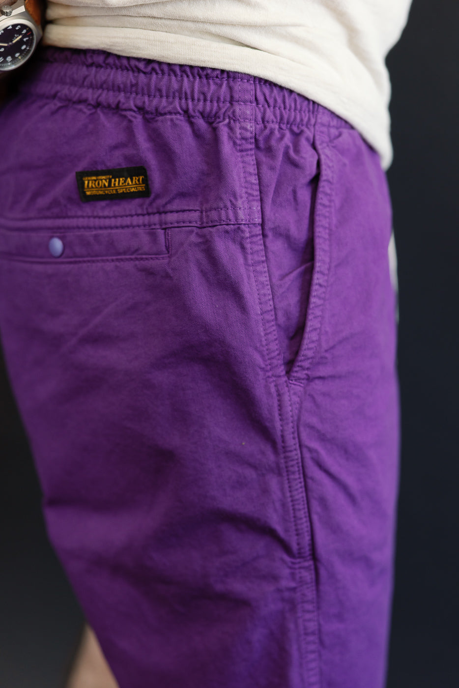 Close-up of male model's side view, showcasing purple shorts with Iron Heart tag, button pocket, and fabric texture.