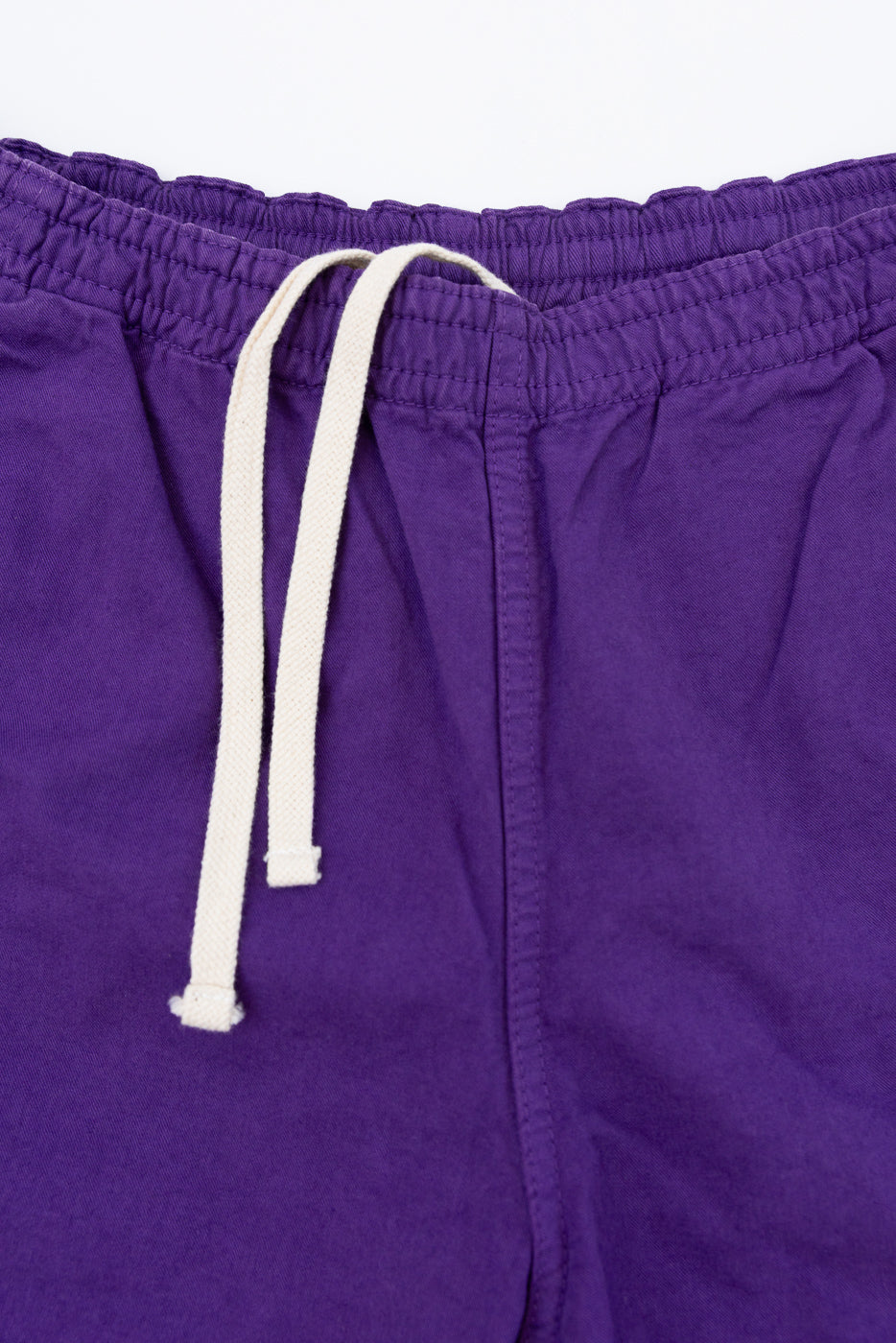 Close-up front view of purple drawstring shorts with cream cord, elastic waistband, and visible stitching detail