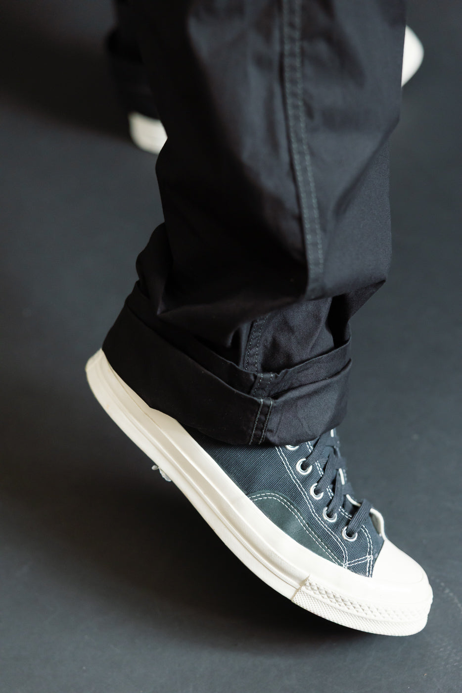 Close-up of male model's lower leg, showcasing black pants with cuffed hem and dark sneakers.