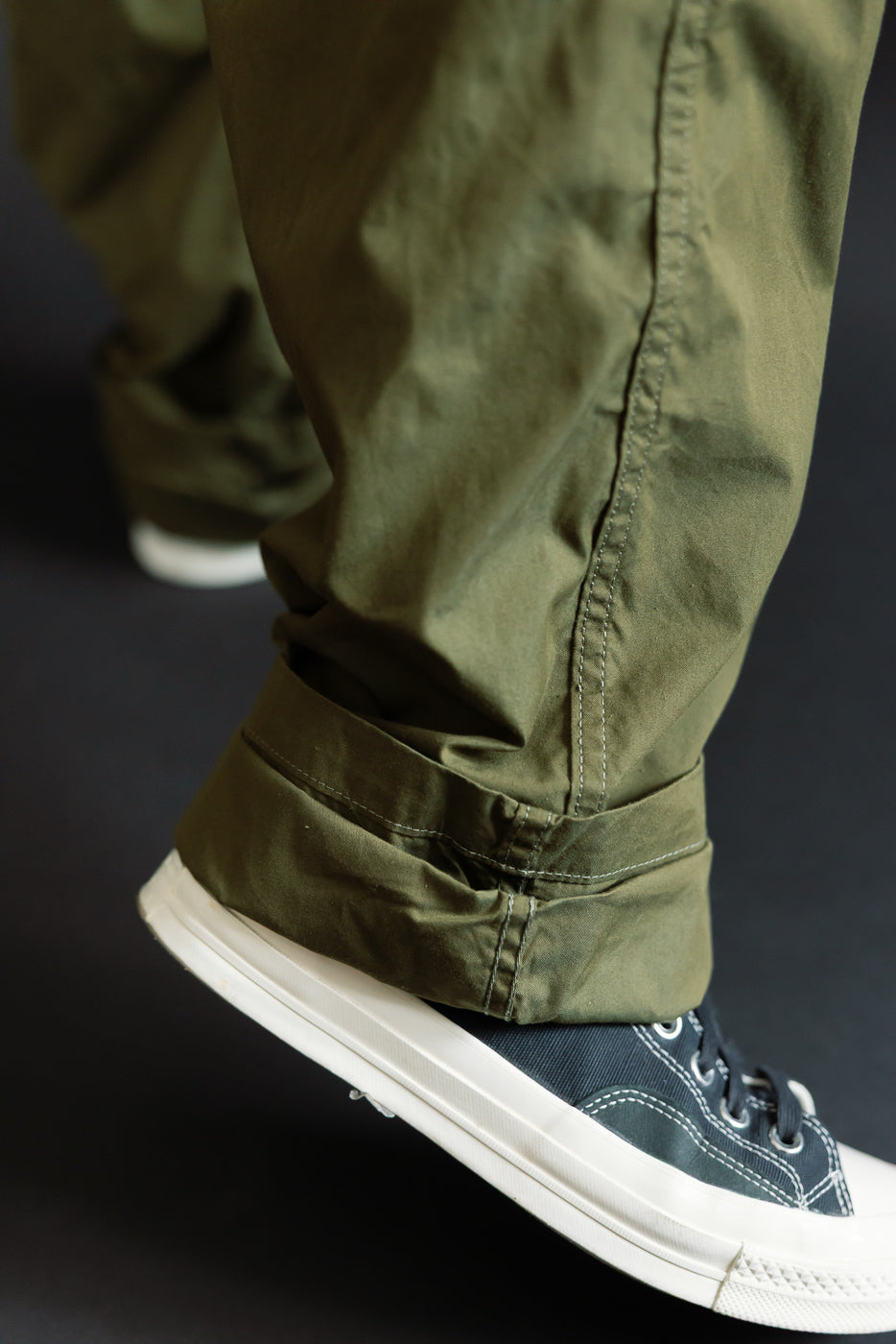 Close-up of olive green pants hem, cuffed, worn by male model with athletic build, paired with navy sneakers