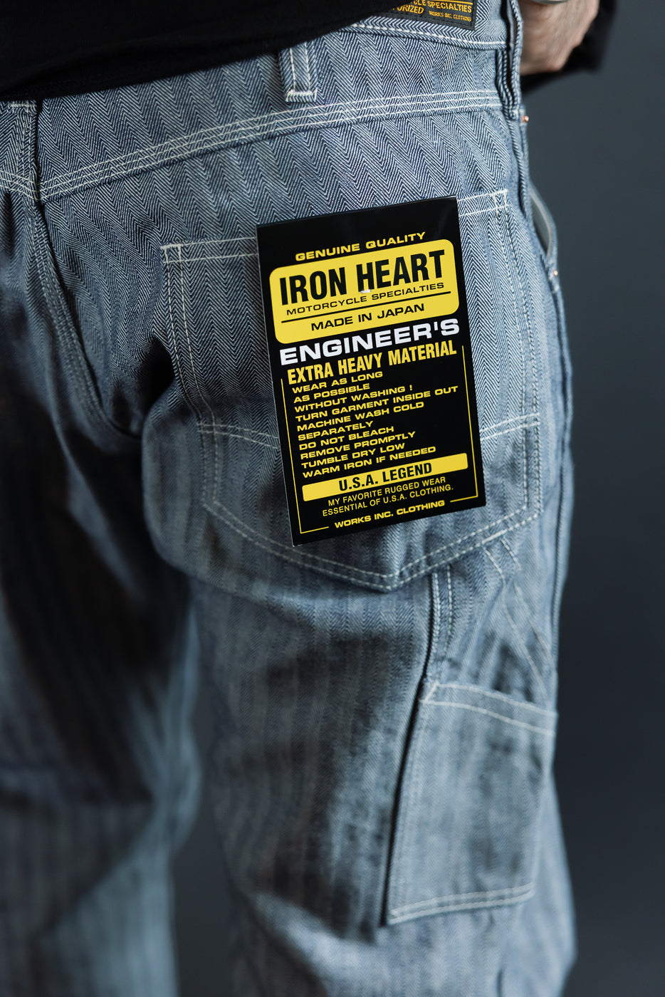 Close-up back view of male model's denim pants showing Iron Heart tag, herringbone texture, and rear pocket detail.