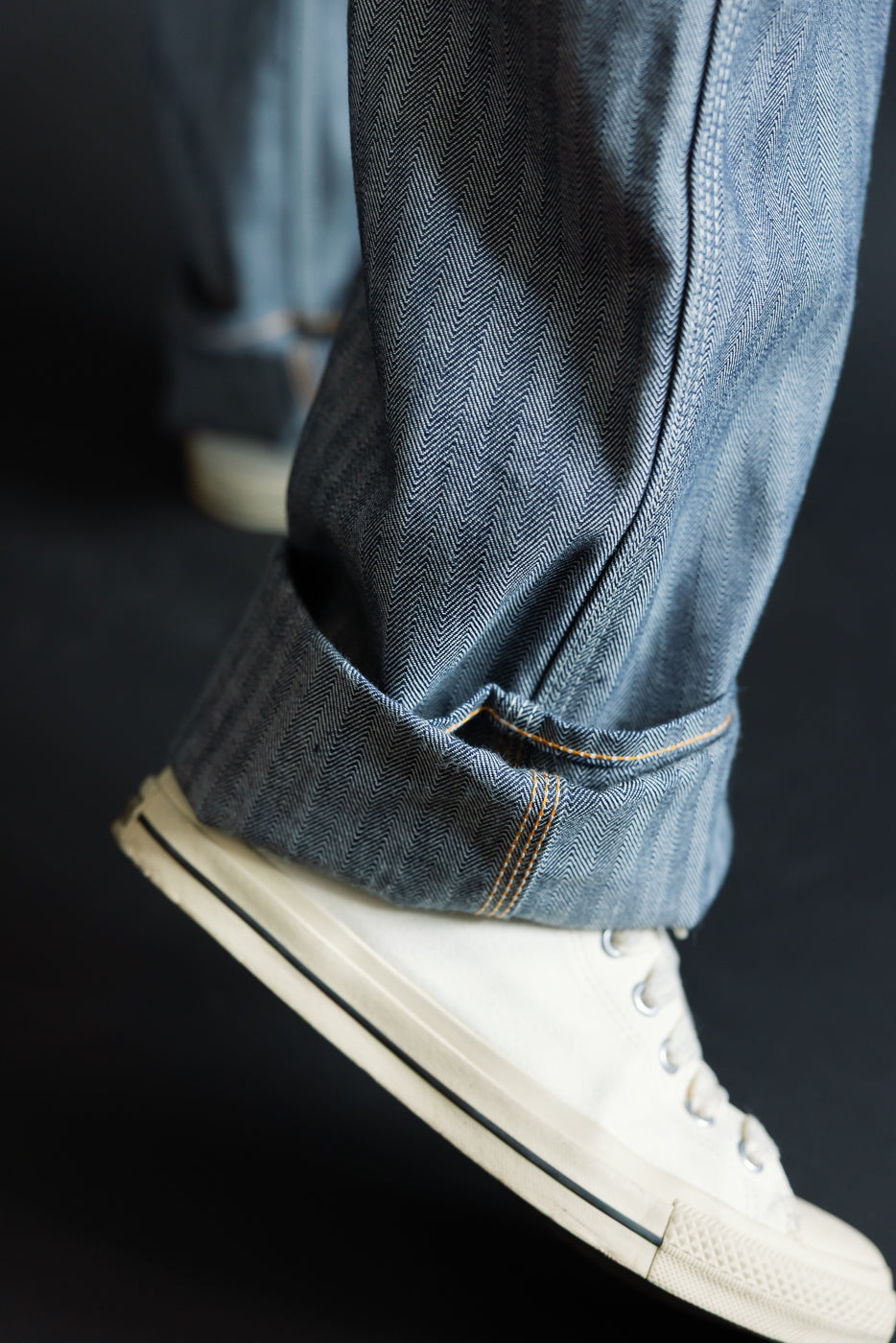 Close-up of male model's leg in herringbone selvedge jeans, cuffed hem with orange stitching, paired with white sneakers