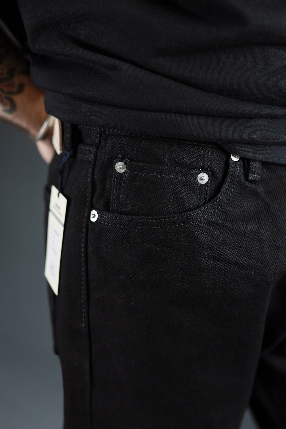 Close-up front view of black selvedge jeans on male model, showing coin pocket, silver rivets, and tag detail